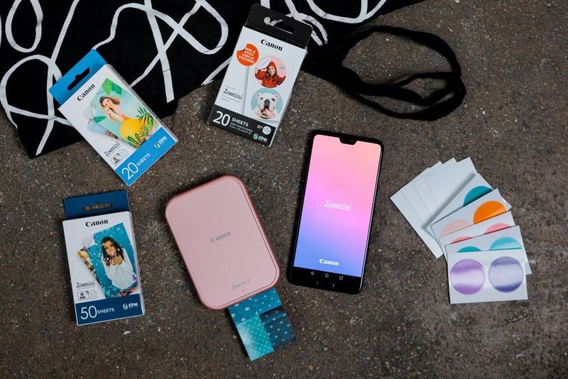  A pink Canon Zoemini portable printer, with a photo emerging from it, sits alongside a smartphone and various packages of media for the printer beside it on the surface.