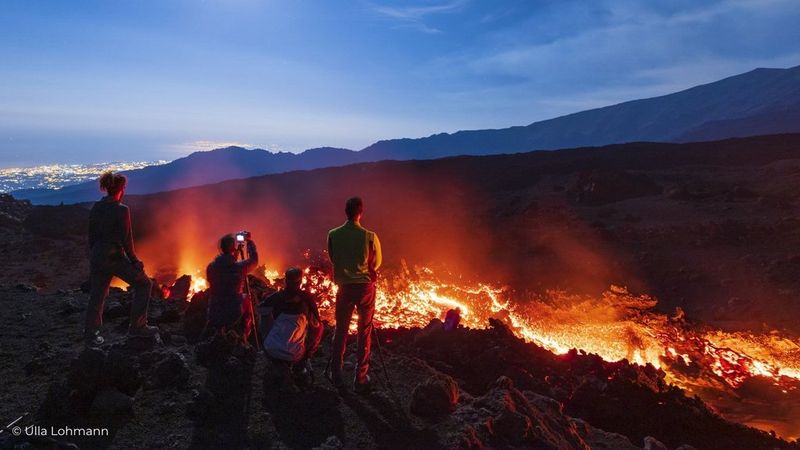 A group of people with cameras stand on a ridge overlooking a glowing lava flow.
