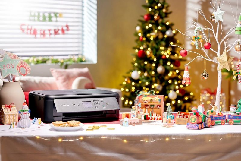 Een Canon PIXMA-printer op een versierde tafel, omringd door zelfgemaakte kerstdecoraties en -ornamenten, met een kerstboom op de achtergrond.