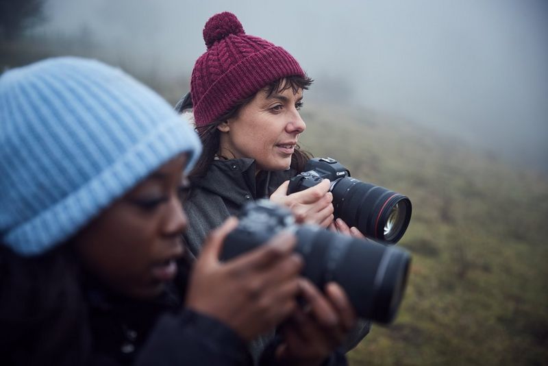 Two women in winter clothing holding Canon cameras stand on a foggy hillside.