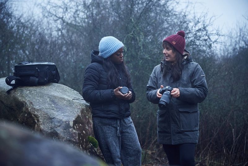Two women in winter clothing talking while they examine camera lenses next to a large rock.