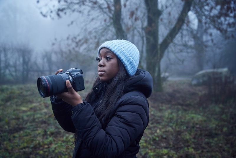 A young woman dressed in winter clothing holds a Canon camera in a foggy wooded area. 