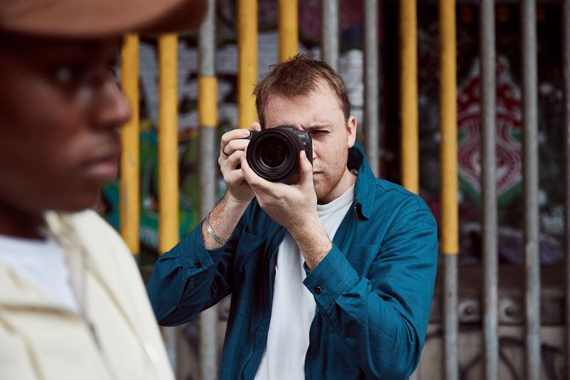 A photographer taking a side profile of a young man in a baseball cap with a Canon EOS RP camera.