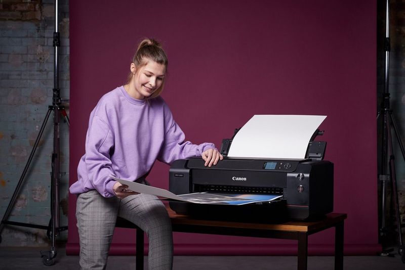 A woman wearing a lilac jumper perches on the edge of a desk holding an A2 print emerging from a Canon imagePROGRAF printer.