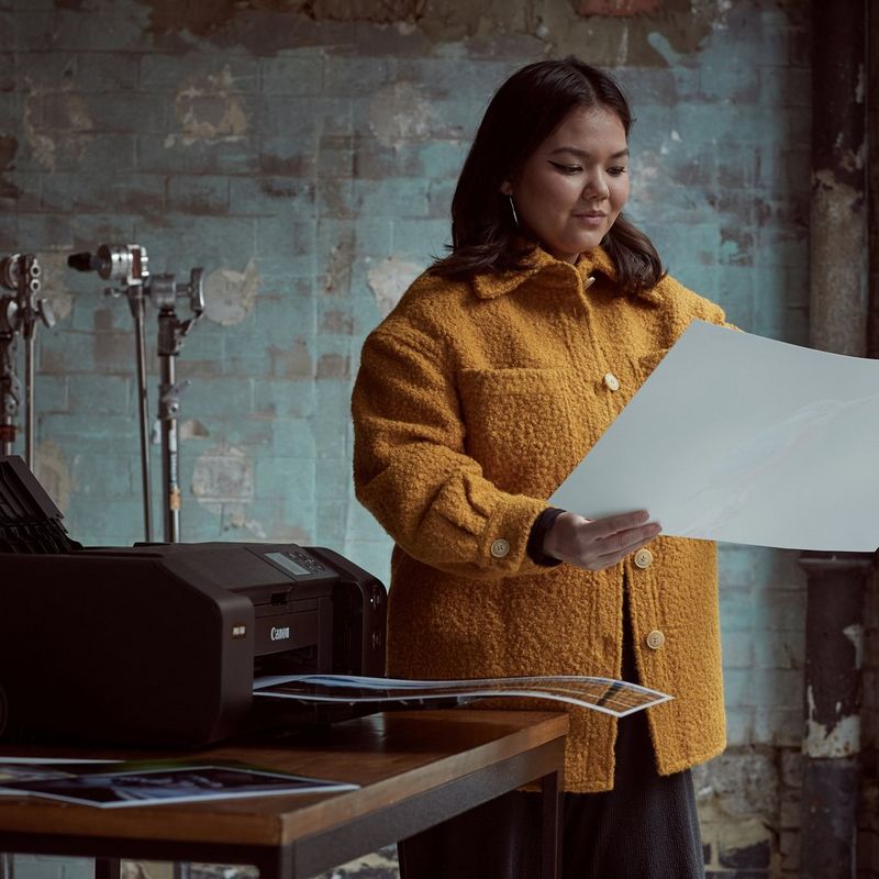 A photography student in a mustard jacket studies an A3 image printed on a Canon imagePROGRAF PRO-300 printer, which is on the table next to her.