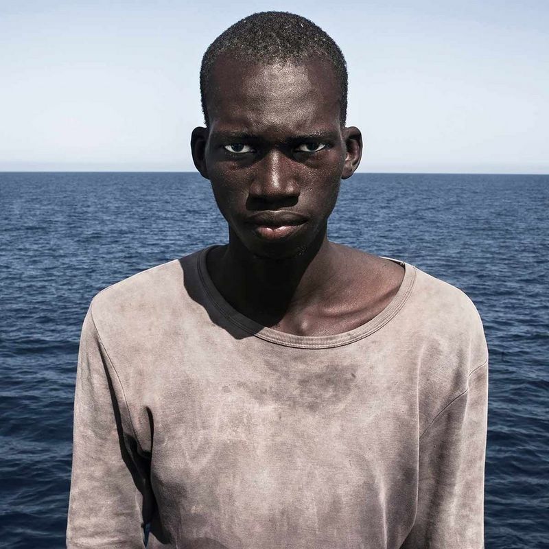 Brows furrowed, 23-year-old migrant Amadou Sumalia stares down the lens, minutes after being rescued from an overcrowded inflatable boat. He is aboard a German NGO vessel, the Iuventa, and the horizon of the Mediterranean can be seen in the backdrop.