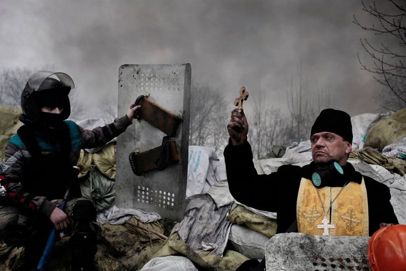 An Orthodox priest blesses protesters along a barricade in Kiev, Ukraine, during protests in 2014, captured by Jérôme Sessini. Unidentified snipers had opened fire on unarmed protesters, with an official sources suggesting 70 people were shot dead. 