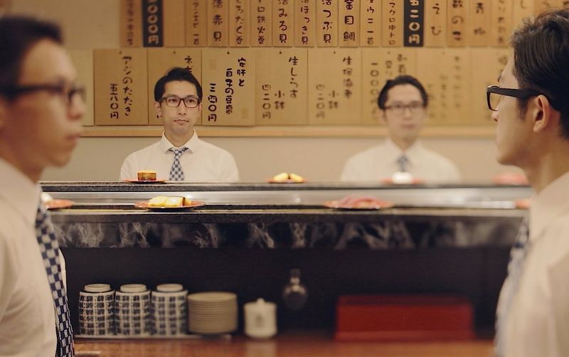 Japanese businessmen sitting in a kaiten-zushi watching sushi plates rotate on a conveyor belt.