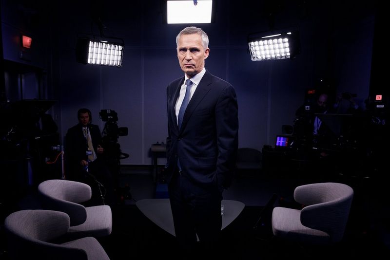 A man in a suit stands in a studio with several chairs, lights, and TV cameras behind him. Shot on Canon.