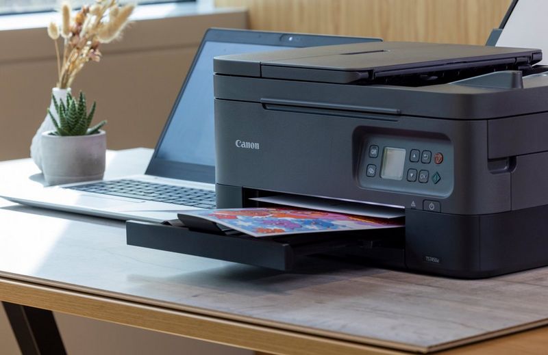 A Canon TS7450a Series printer prints a sheet of paper with bright colours on it. A laptop sits beside the printer on a desktop.