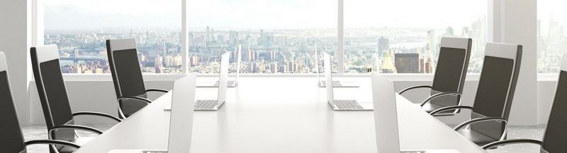 A boardroom table with six chairs and six laptops, against a backdrop of a window, overlooking a city. 
