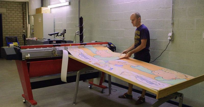 Bald man in purple t-shirt with yellow writing pulls large sheets of multi-colored printed paper from a digital printer.