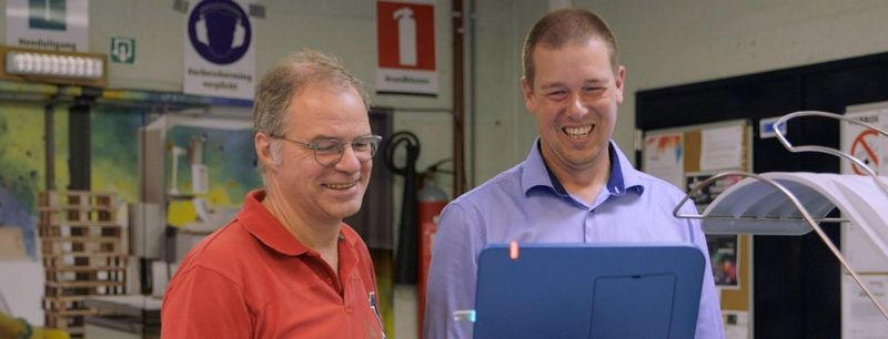 Older man wearing glasses and an orange polo shirt and younger man in blue long-sleeved shirt smile as they look at a screen on a commercial digital printer.