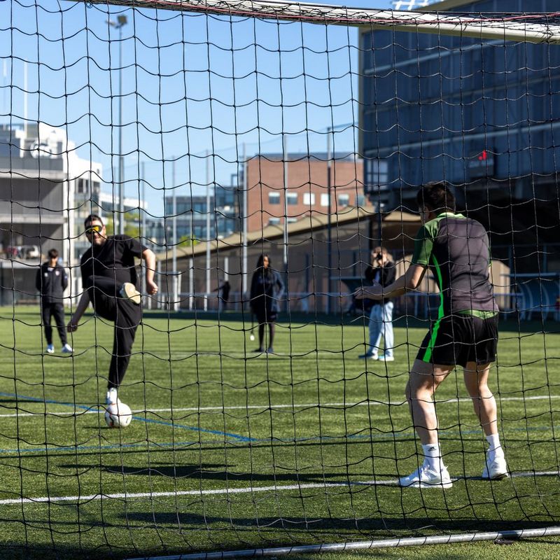 A football player in an eye mask and black kit kicks a ball towards the goalkeeper on an outdoor turf field, viewed through the mesh of a goal net.