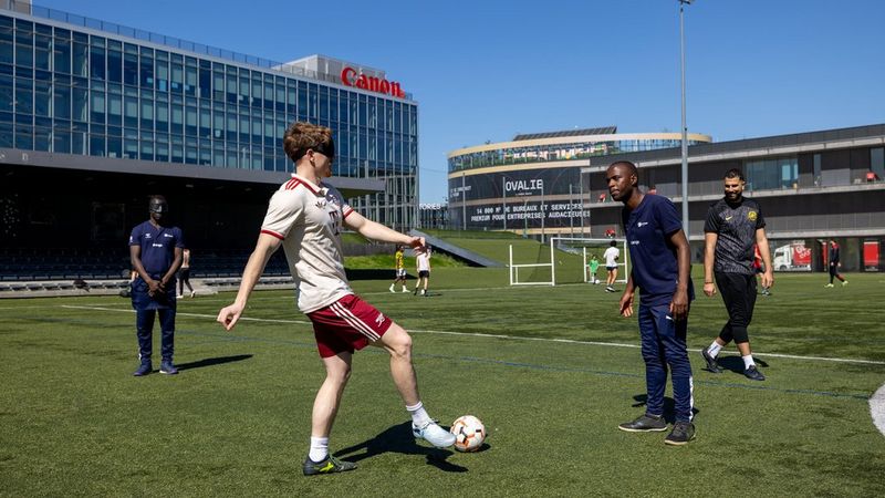 A cecifoot player in an eye mask, wearing a white and red kit dribbles a ball on a green pitch, guided by a coach standing nearby under a clear blue sky.