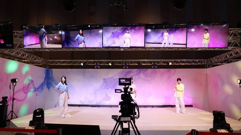 Three young people stand on a stage in front of a camera, beneath a bank of five screens showing different areas of the stage.