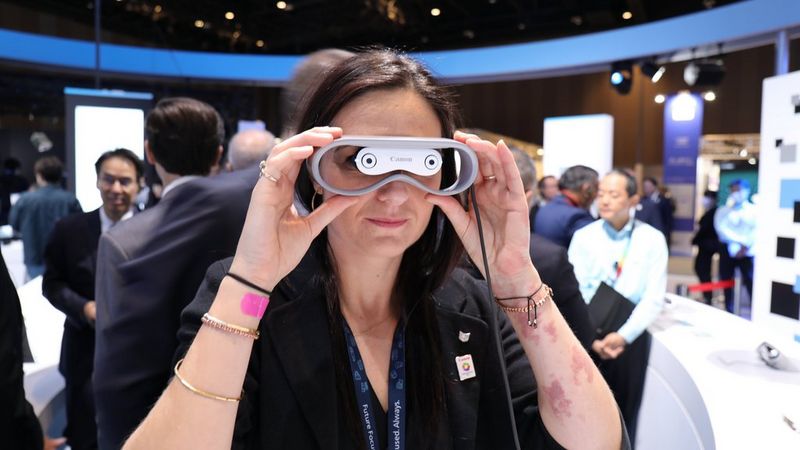 A woman with long dark hair peers through a pair of handheld AR glasses.