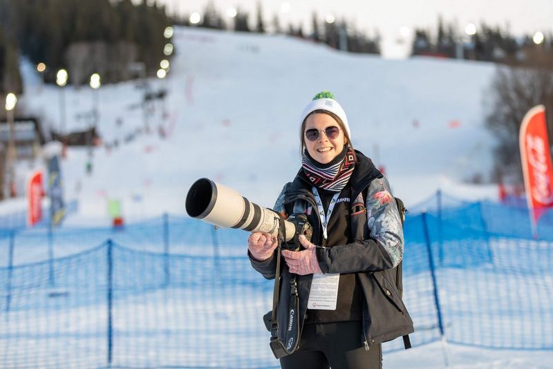 A photograph of Claire Behan, against a backdrop of a snow-covered winter sports course. She is wearing a white and green wool hat that covers her ears and sunglasses, as well as a striped scarf and Canon-branded gilet. In her hand she holds a Canon camera with a huge L-series lens and has an accreditation pass on a lanyard around her neck.