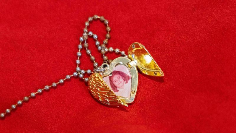 A gold, heart-shaped locket lies on a background of a blood red fabric. The front opening of the locket is two wings meeting and one wing is open, revealing an old, fading photograph of a little girl.