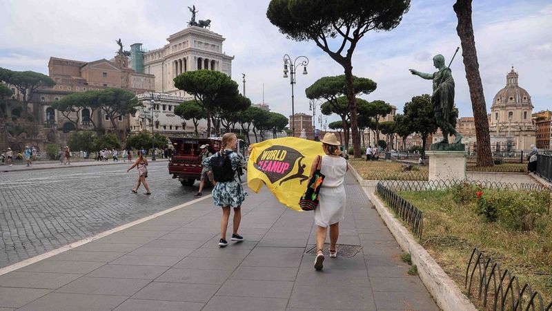 Two women walk through the streets of Rome holding a yellow flag between them. The flag reads "World Cleanup"
