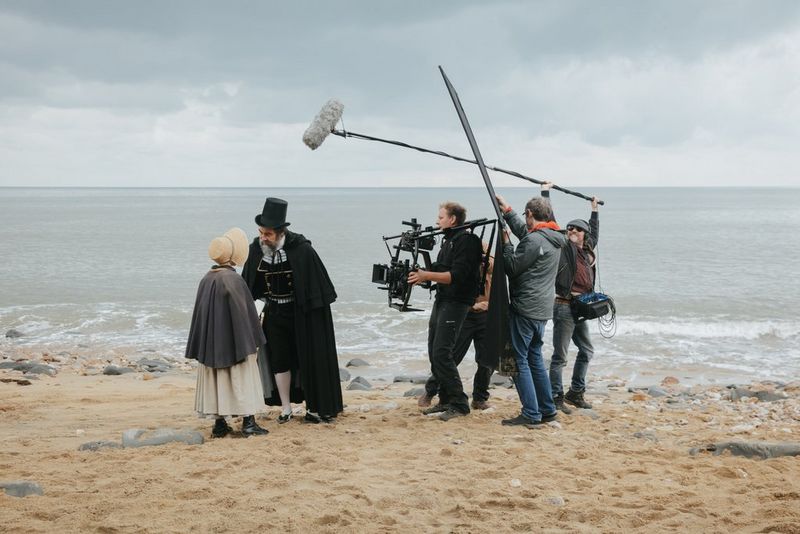 A camera crew filming two actors beside the sea: a Victorian gentleman in a top hat and cape leaning over to talk to a young girl.