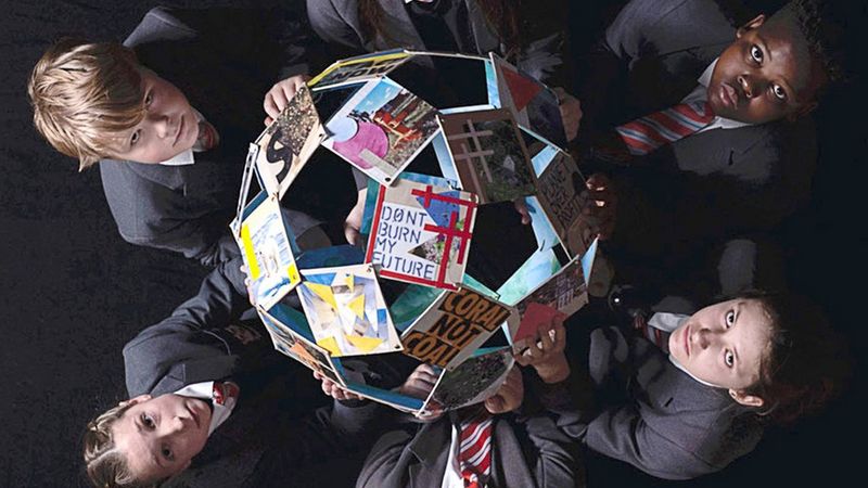 Taken from above, two children, on the left and right of the photo, hold a globe made of photographs and raise their solemn faces to the camera. The clearest photo, closest to the camera displays the words ‘don’t burn my future’."
