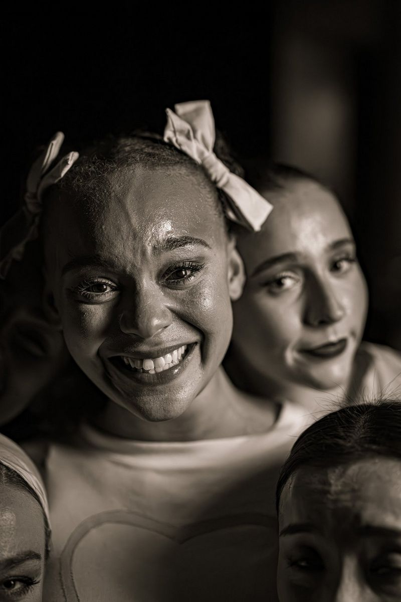 Een zwart-witfoto van een zwetende, lachende jonge vrouw met strikjes in haar haar, die rechtstreeks naar de camera kijkt. Drie andere jonge vrouwen zijn gedeeltelijk zichtbaar op de achtergrond.