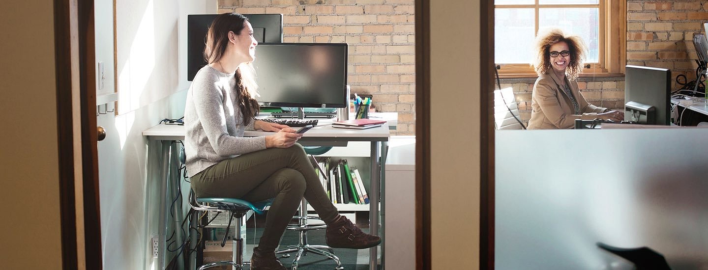 Woman holding a mobile phone and sitting cross-legged at a desk next to a PC chats to a colleague across the office.