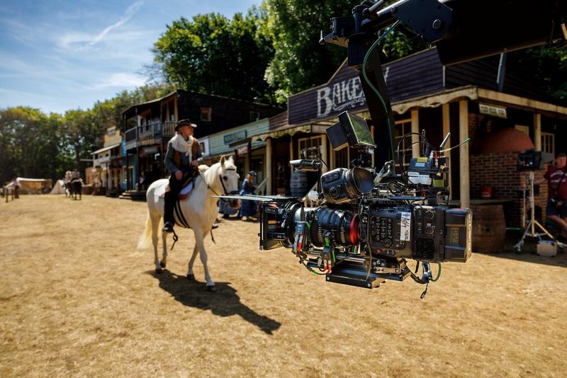 A large camera rig is filming a man on a white horse riding towards the camera, in the street of a Wild West town.