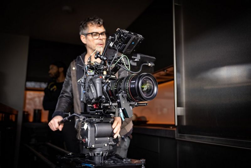 A man with dark hair and glasses stands operating a large camera which is set up on a stand.