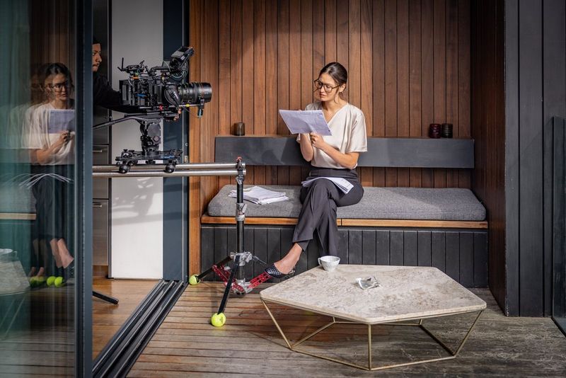A woman sits cross-legged on a padded bench, reading some papers. To the side of her is a large camera filming her at head height.