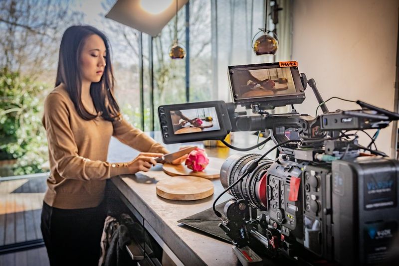 A woman stands at a kitchen worktop chopping fruit. She is being filmed by a large camera that is sitting on the worktop in front of her.