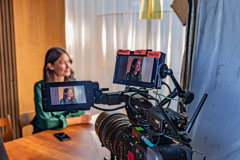 A woman sitting at a table is being filmed by a large camera, with her image being shown on two viewscreens above the camera.