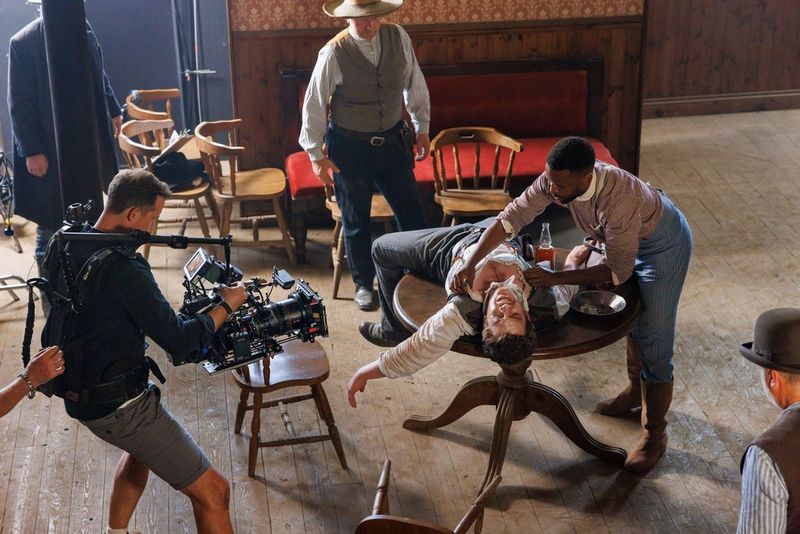 A man lies on his back across a round table in a Western bar, looking injured. Another man leans over him, pulling open his shirt. Next to them a cameraman films the scene on a large camera and lens.