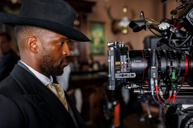 A man wearing a suit and black hat stares straight into the lens of a large camera rig.