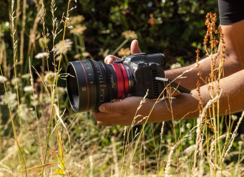 In a field of grass, a pair of hands holds a Canon EOS R5 C with a CN-R Prime lens attached to it.