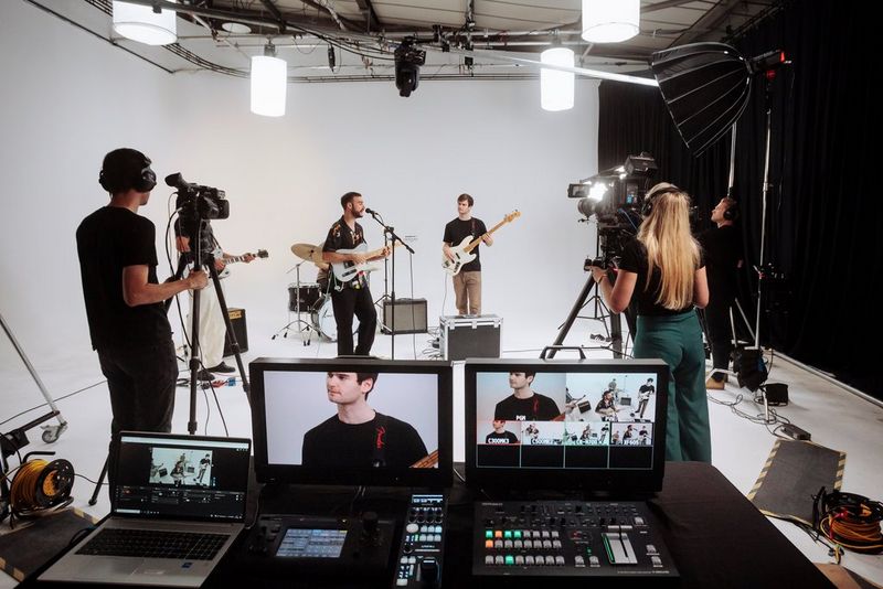 Een crew filmt een band die optreedt op een witte set. De bandleden zijn zichtbaar op de twee schermen op de voorgrond.