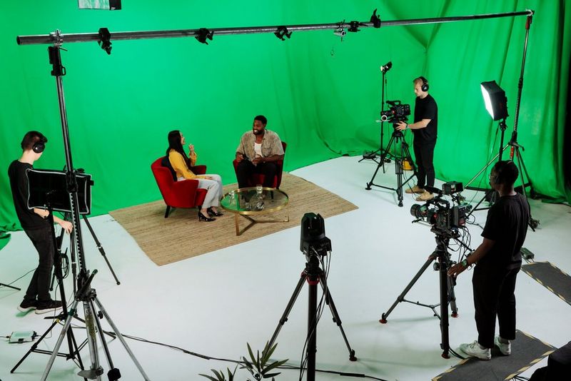 A video production crew films two people seated on a studio set surrounded by green screens, cameras and professional lighting equipment.