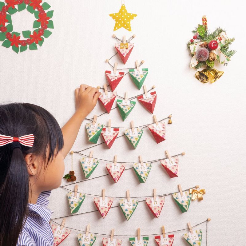 Een jong kind reikt naar een envelop op een zelfgemaakte adventskalender in de vorm van een kerstboom en gemaakt van rode en groene papieren enveloppen. De enveloppen hangen aan koorden aan de wand. Voor dit project is een Canon Creative Park-sjabloon voor PIXMA-printers gebruikt.