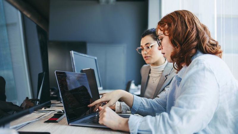 Two women sit side by side at a desk with laptops in front of them. One points at a screen and they appear to discuss what they are seeing.