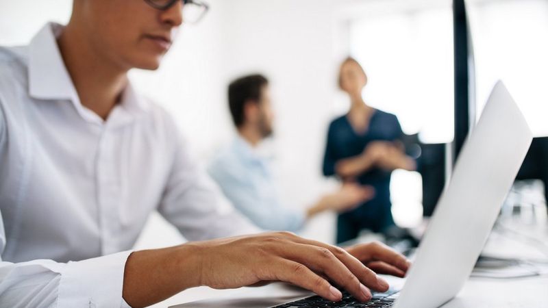 Lightly blurred image of a man in a white shirt, typing on a laptop. In the background are the figures of two people talking.