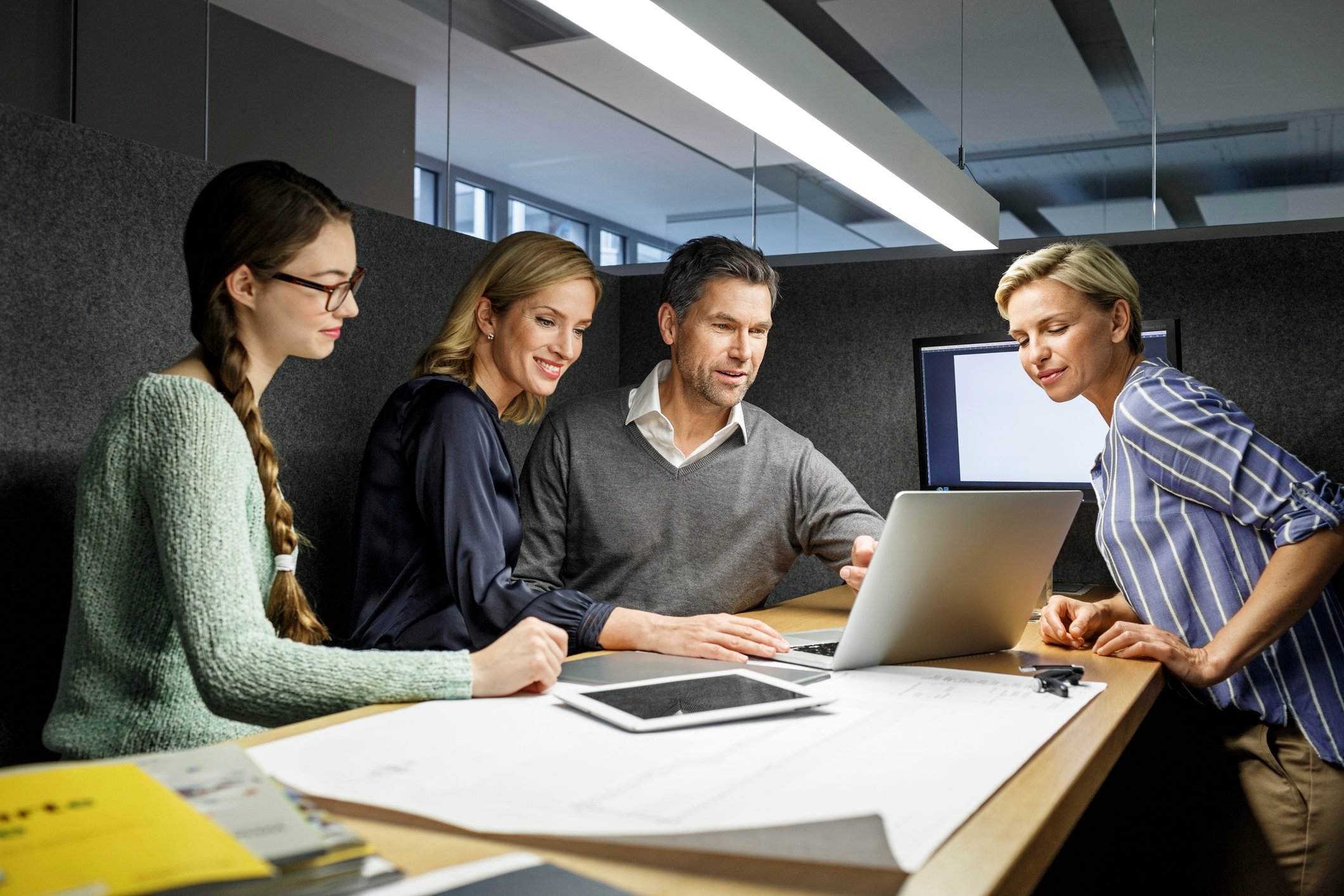 Three women and a man dressed in business casual sit around a table in a partitioned office, looking at a laptop screen.