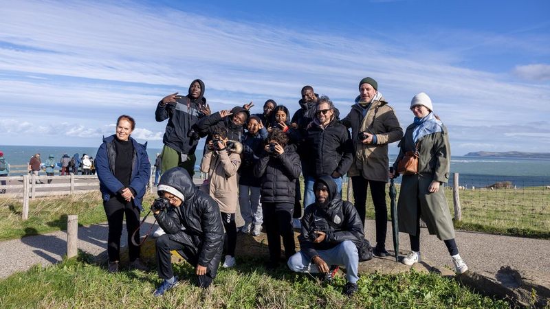 Een groep jongeren, waarvan een aantal met een camera in de hand, poseert samen aan de kust onder een blauwe lucht.