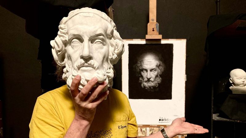 A man in a yellow t-shirt stands holding a white sculpture of the head of a bearded man in front of his own face, in his right hand. His left-hand gestures, open-palmed, towards a drawing on an easel behind him. It is a representation of the sculpted head in charcoal, on a black background.