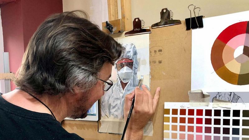 Photographed from behind, a man in a black t-shirt stands in front of a board upon which a painting of a person dressed in full PPE is taped. Next to it is a colour wheel and colour chart, also taped onto the board.