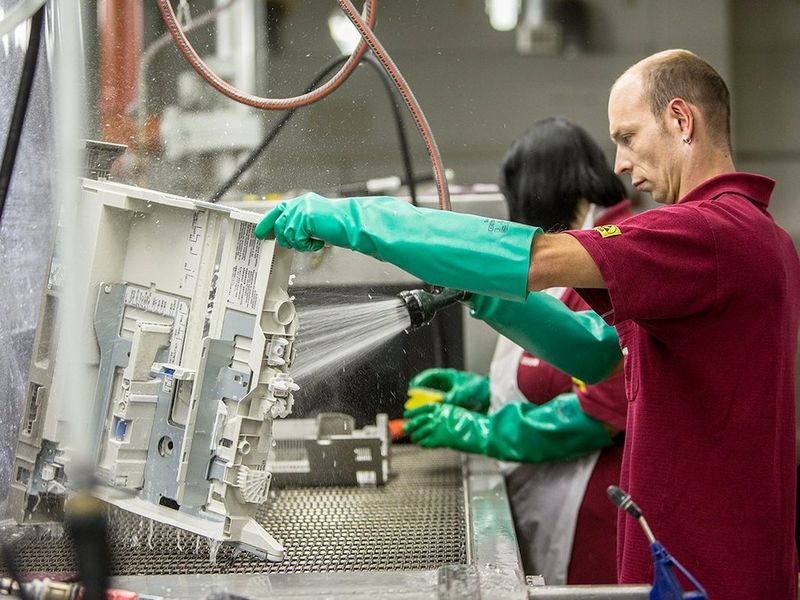 A man in a deep red shirt and green, elbow length rubber gloves, is in an industrial setting and cleaning a section of a machine using a powerful water spray.