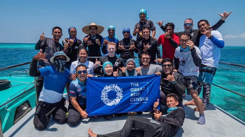 A smiling group of conservationists pose on a boat deck. The hold a blue banner that reads ‘The Great Reef Census’.