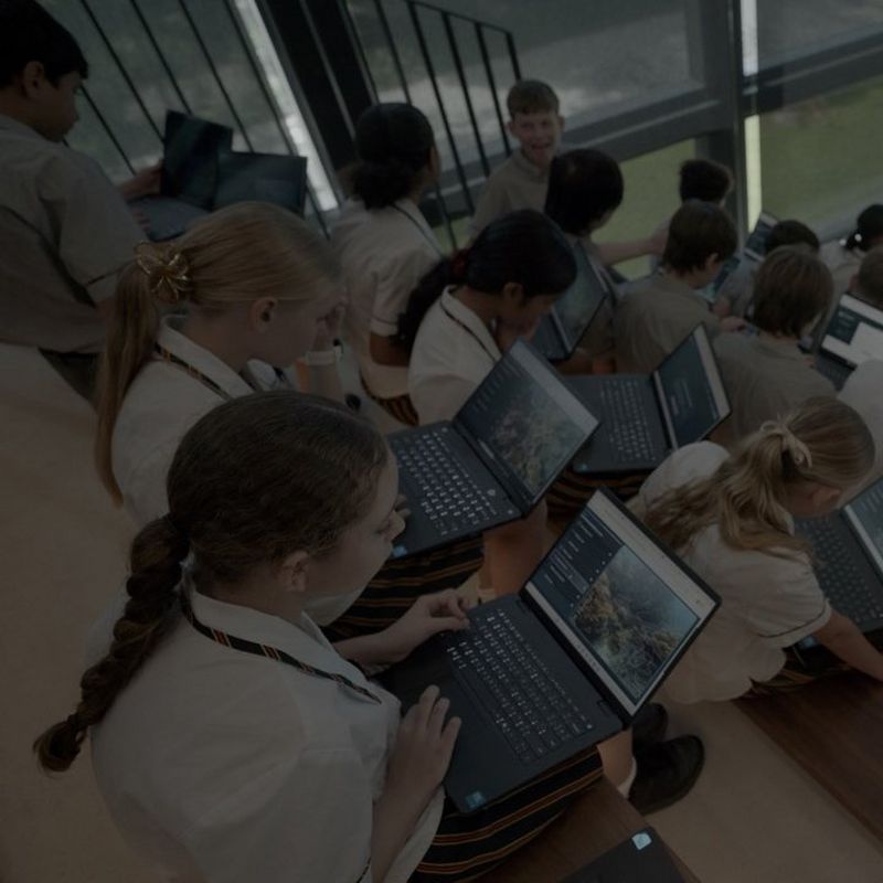Students in school uniforms sit on tiered seating, working on laptops displaying coral reef imagery.