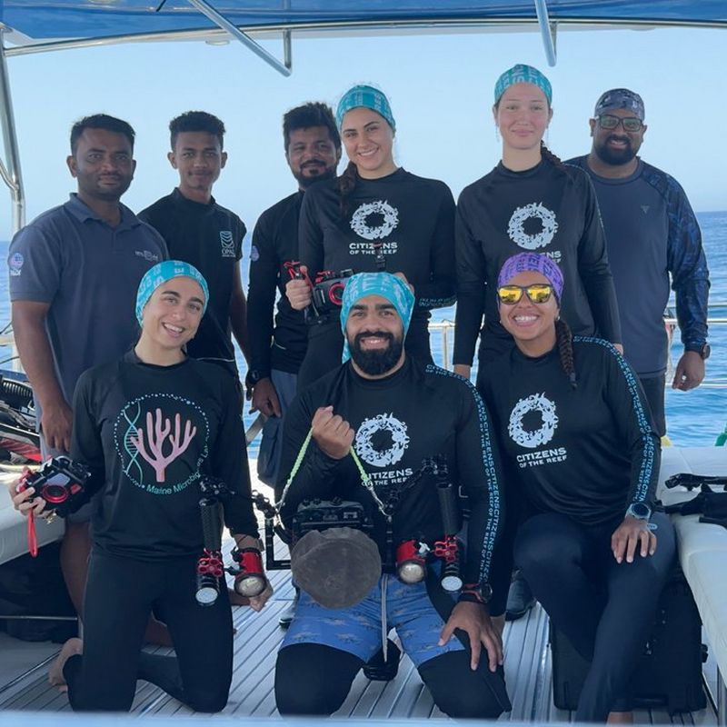 A team of divers wearing matching Citizens of the Reef shirts pose together with underwater camera gear on a boat.