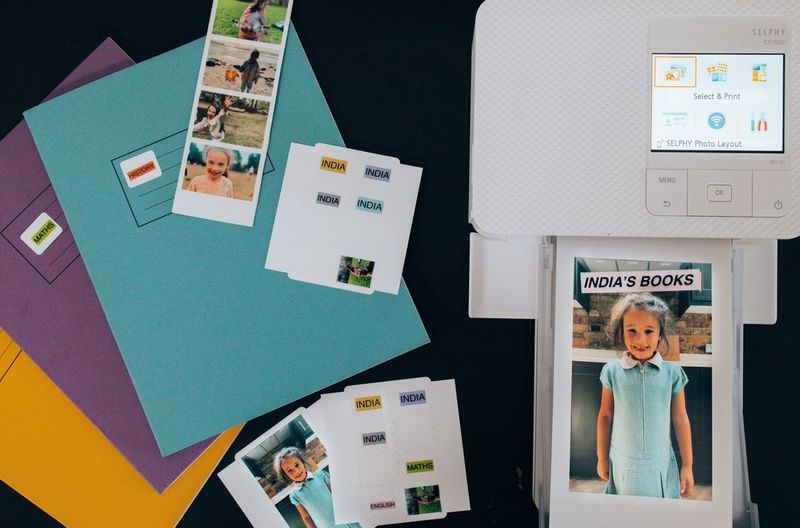 A top-down image of colourful exercise books, sheets of stickers and a Canon SELPHY CP1500 printing a picture of a young girl in school uniform.